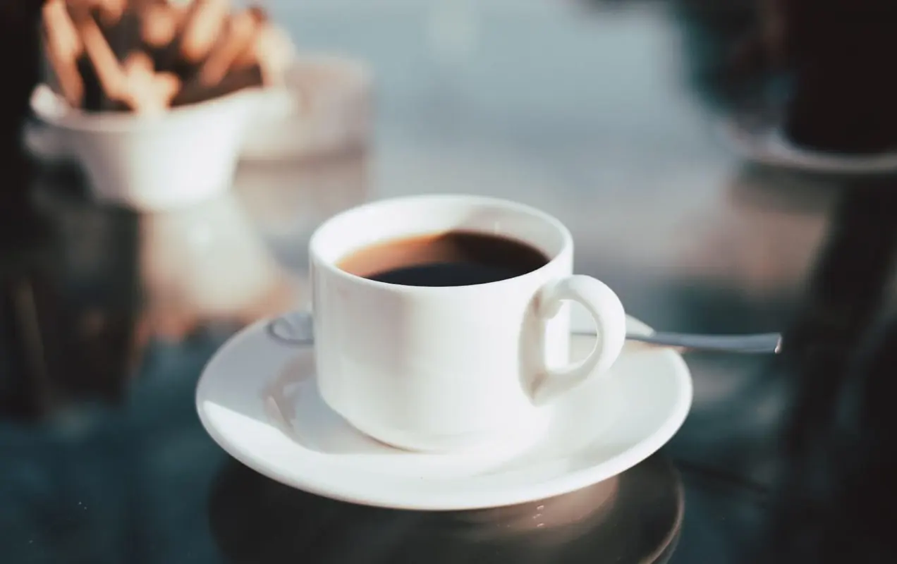 Uma mesa de vidro reflexiva com uma xícara de café branca curta cheia de café preto, sentada em um pires branco