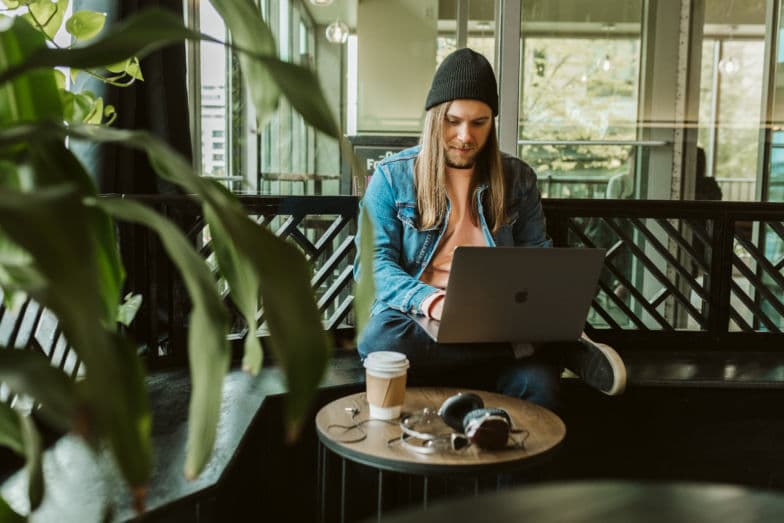 Ein Mann arbeitet an einem Laptop, während er vor einem kleinen Tisch sitzt, auf dem Kaffee und Kopfhörer stehen