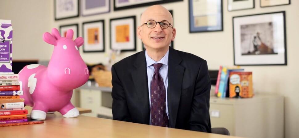 La imagen de Seth Godin donde se sienta en su mesa de lectura con un unicornio y muchos libros a su alrededor.