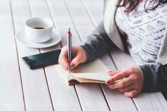 une femme écrit dans un cahier sur un bureau blanc à côté de son téléphone et d'une tasse de café à moitié pleine