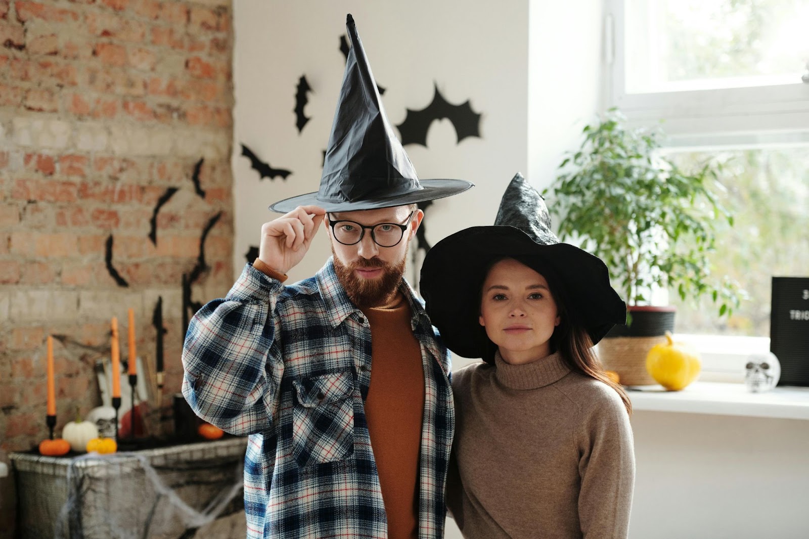 Um homem e uma mulher usando chapéus de bruxa em um escritório rústico decorado com outros elementos sazonais, como velas, abóboras e caveiras.